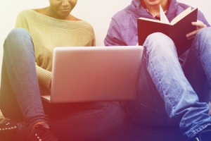 Two college students, one female, the other male, are sitting against a wall; in her lap she is working on a laptop; the male student is reading from a book. Their faces are mostly out of frame.