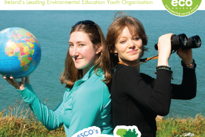 Two female students sitting back to back faces turned to camera smiling, one with binoculars in hand, the other with a small globe. In the background, the sea is visible. The magazine title is ECO-UNESCO, Ireland's leading environmental education youth organisation
